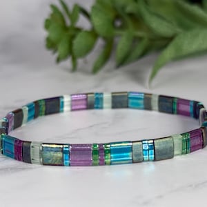 May include: A colorful bracelet with rectangular beads in shades of teal, purple, green, and silver. The bracelet is made of small, flat beads strung together to form a flexible band. The background is a blurred white surface with a green plant.