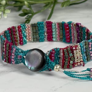 May include: A beaded bracelet with a turquoise base, featuring rows of red, pink, and green beads. The bracelet has a large, round, iridescent button clasp and a tassel with small beads.