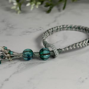 May include: A braided light gray cord bracelet featuring two large, ribbed, teal and dark blue glass beads. The ends of the cord are finished with a tassel containing smaller metallic silver beads and tiny teal glass seed beads.