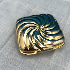 May include: A square gold-toned compact case featuring a raised, swirling sunburst pattern on the lid. The metallic surface has a reflective finish with deep blue and dark green shadows accentuating the curved, radiating grooves of the design.
