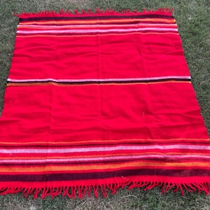 May include: A rectangular woven blanket featuring a vibrant red base color with horizontal stripes in shades of orange, yellow, pink, and dark brown. The blanket has a textured surface and is finished with fringe along the top and bottom edges.