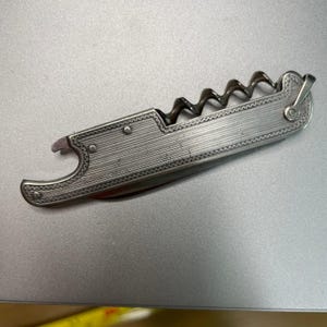 May include: A silver-toned metal multi-tool featuring a corkscrew, a bottle opener, and a folding blade. The handle has a textured, linear engraved pattern with a decorative border and small rivets along the side of the frame.