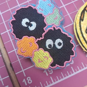 May include: Embroidered patches featuring cartoon-like characters with black bodies, white eyes, and colorful accents. The patches have a light blue border and include orange, yellow, and blue details. The patches are on a pink cutting mat.
