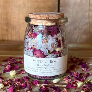 May include: A clear glass jar filled with pink rose petals, white crystals, and dried botanicals. The jar has a cork lid and a label that reads "VINTAGE ROSE floral bath tea". The jar is surrounded by loose petals and botanicals.