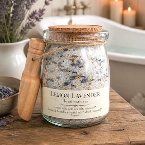May include: A clear glass jar filled with lemon lavender floral bath tea, featuring a cork lid and a wooden scoop tied with twine. The label reads: LEMON LAVENDER floral bath tea, epsom salt, dead sea salt, jojoba oil, lemon & lavender essential oils, dried botanicals, organic.