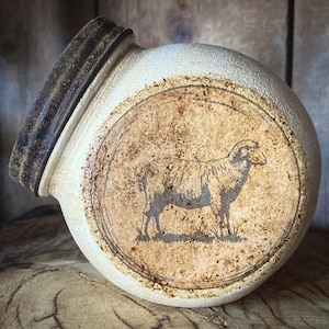 May include: A cream-colored ceramic jar with a dark brown rim and a circular design featuring a sheep. The jar has a rustic, aged appearance, with a textured surface and a slightly tilted angle. The sheep illustration is in a brown tone.