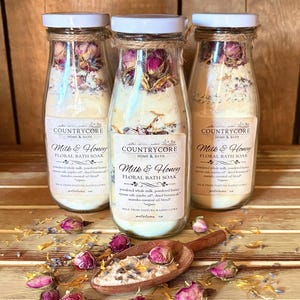 May include: Three glass bottles filled with Milk & Honey Floral Bath Soak, featuring dried rosebuds and botanicals. Each bottle has a white lid and a label reading Countrycore Home & Bath. A wooden scoop contains the bath soak mixture.