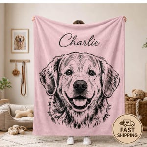 Puede incluir: Una manta de forro polar rosa claro suave que presenta una gran ilustraci&oacute;n detallada en l&iacute;neas negras del rostro de un golden retriever. El nombre Charlie est&aacute; impreso en una tipograf&iacute;a cursiva negra sobre la ilustraci&oacute;n del perro. La manta es rectangular.