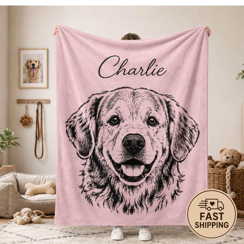 Puede incluir: Una manta de forro polar rosa claro suave que presenta una gran ilustraci&oacute;n detallada en l&iacute;neas negras del rostro de un golden retriever. El nombre Charlie est&aacute; impreso en una tipograf&iacute;a cursiva negra sobre la ilustraci&oacute;n del perro. La manta es rectangular.