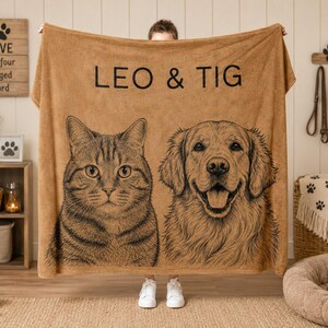 Puede incluir: Una manta de forro polar color canela con una ilustraci&oacute;n de l&iacute;neas negras de un gato atigrado y un golden retriever. El texto LEO & TIG est&aacute; impreso en letras may&uacute;sculas negras sobre los retratos de los animales en la tela rectangular.