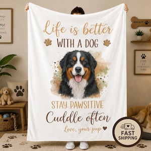 May include: A white fleece blanket featuring a watercolor illustration of a Bernese Mountain Dog. The text reads: Life is better WITH A DOG, STAY PAWSITIVE, Cuddle often, Love, your pup. The design includes brown paw prints and a small heart icon.