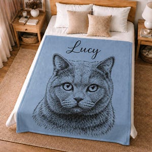 Puede incluir: Una manta de forro polar azul claro con una detallada ilustraci&oacute;n de l&iacute;neas negras de una cara de gato centrada en la tela. El nombre Lucy est&aacute; impreso en una fuente cursiva negra sobre la ilustraci&oacute;n del gato. La manta est&aacute; extendida.