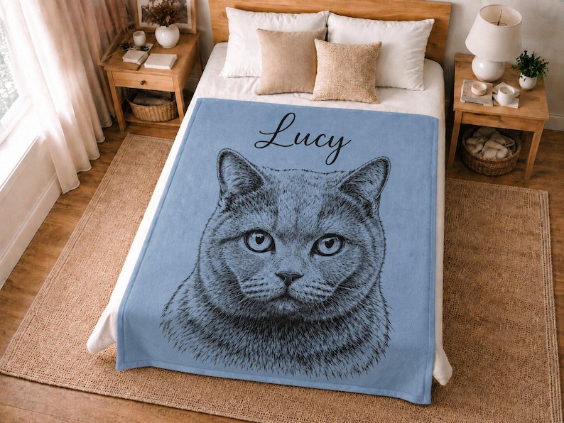 Puede incluir: Una manta de forro polar azul claro con una detallada ilustraci&oacute;n de l&iacute;neas negras de una cara de gato centrada en la tela. El nombre Lucy est&aacute; impreso en una fuente cursiva negra sobre la ilustraci&oacute;n del gato. La manta est&aacute; extendida.