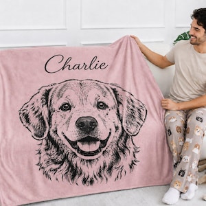 Puede incluir: Una manta de forro polar de color rosa empolvado suave con una ilustraci&oacute;n de l&iacute;neas negras de la cara de un golden retriever en el centro. El nombre Charlie est&aacute; impreso en cursiva negra sobre la ilustraci&oacute;n del perro.