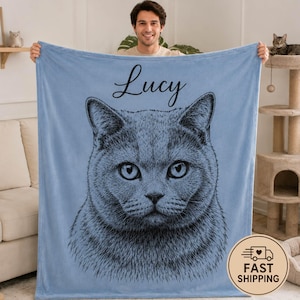 Puede incluir: Una manta de forro polar azul claro con una ilustraci&oacute;n detallada de l&iacute;neas negras de la cara de un gato centrada en la tela. Sobre la ilustraci&oacute;n del gato, el nombre Lucy est&aacute; impreso en una fuente cursiva negra en la parte superior.