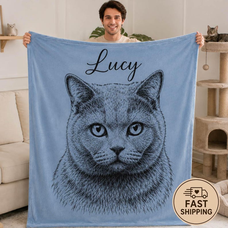 Puede incluir: Una manta de forro polar azul claro con una ilustraci&oacute;n detallada de l&iacute;neas negras de la cara de un gato centrada en la tela. Sobre la ilustraci&oacute;n del gato, el nombre Lucy est&aacute; impreso en una fuente cursiva negra en la parte superior.