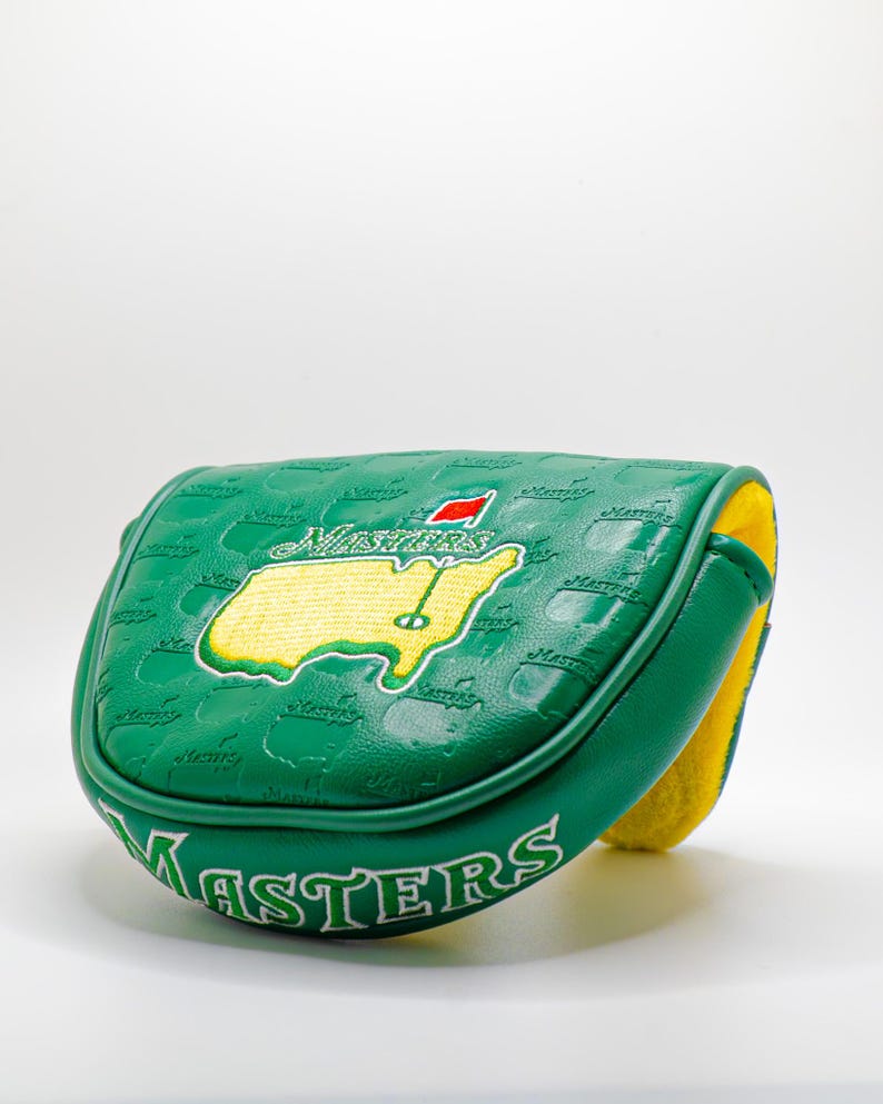 May include: Green golf club head cover with the Masters logo. The cover features a map of the course in yellow and green, with a red flag. The word "Masters" is printed on the cover.