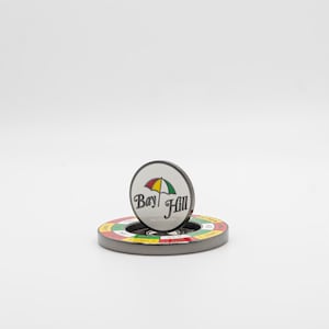 May include: A golf ball marker with the words "Bay Hill" and a colorful umbrella logo. The marker is resting on a circular base with a colorful design. The background is plain white.
