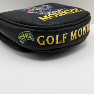 May include: A black leather golf putter headcover featuring embroidered yellow text that reads GOLF MONKEY. The design includes a colorful embroidered graphic of a monkey and a green dollar bill icon on the side of the black textured material.