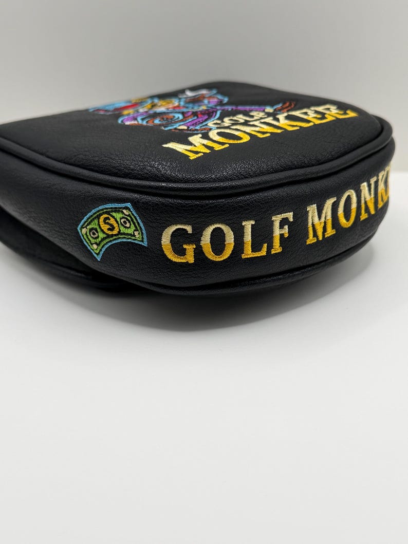 May include: A black leather golf putter headcover featuring embroidered yellow text that reads GOLF MONKEY. The design includes a colorful embroidered graphic of a monkey and a green dollar bill icon on the side of the black textured material.
