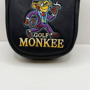 May include: A black leather-like golf putter headcover featuring an embroidered graphic of a monkey wearing a top hat, monocle, and suit, holding a cigar. The text GOLF MONKEE is embroidered in yellow below the monkey figure on the front surface.
