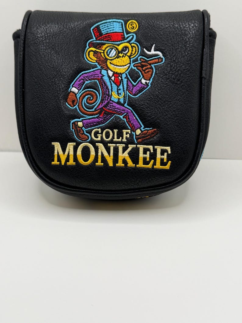 May include: A black leather-like golf putter headcover featuring an embroidered graphic of a monkey wearing a top hat, monocle, and suit, holding a cigar. The text GOLF MONKEE is embroidered in yellow below the monkey figure on the front surface.