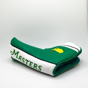 Puede incluir: Funda para la cabeza del palo de golf verde y blanca con la palabra "MASTERS" en escritura verde. La funda presenta un logotipo verde y amarillo y rayas blancas y negras.