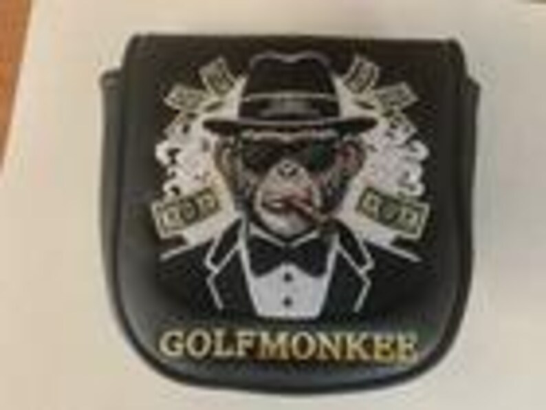 May include: A black golf putter headcover featuring a graphic of a monkey wearing a tuxedo, fedora, and sunglasses while smoking a cigar. The design includes floating dollar bills and the text GOLFMONKEE printed in gold at the bottom.