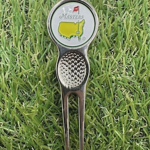 May include: A silver golf divot tool with a ball marker featuring the Masters logo. The marker has a green outline, a yellow map of Georgia, and the word "Masters" above it. The tool is on a bed of green grass.