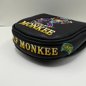 May include: A black leather-like golf putter headcover featuring embroidered yellow text that reads GOLF MONKEE. The design includes a colorful embroidered graphic of a monkey wearing a hat and suit, along with a small green dollar bill icon.