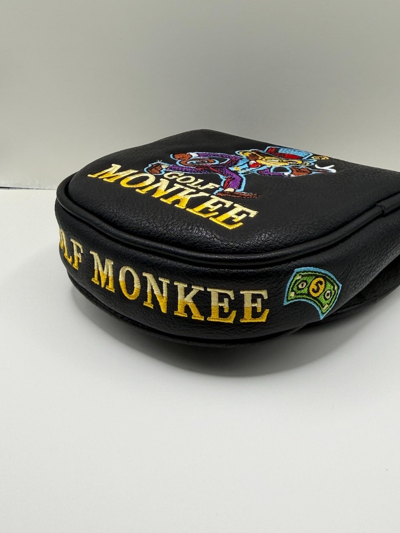 May include: A black leather-like golf putter headcover featuring embroidered yellow text that reads GOLF MONKEE. The design includes a colorful embroidered graphic of a monkey wearing a hat and suit, along with a small green dollar bill icon.