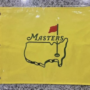 Peut inclure: Drapeau de golf jaune avec le logo Masters. Le logo comprend le mot "Masters" en vert au-dessus d'un contour vert d'une carte avec un drapeau rouge et un trou de golf. Le drapeau a trois œillets.