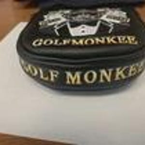 May include: A black golf putter headcover featuring white embroidered text that reads GOLFMONKEE on the top and side. The top design includes a white graphic of a tuxedo jacket with a bowtie. The cover has a rounded shape and stitched edges.