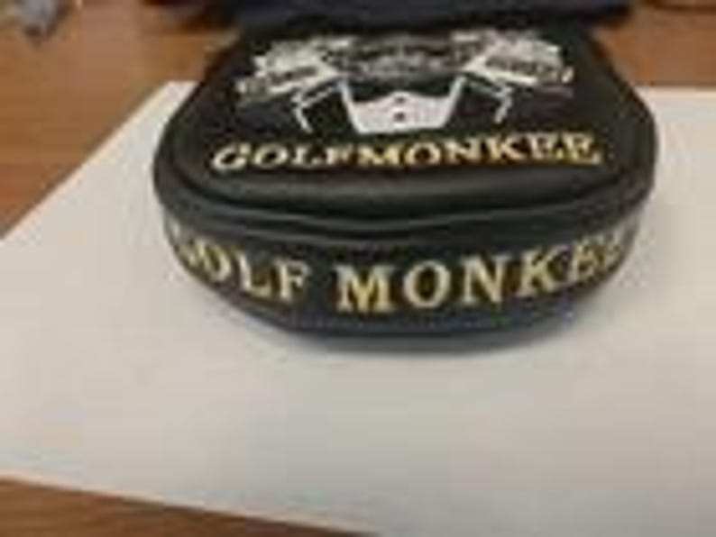 May include: A black golf putter headcover featuring white embroidered text that reads GOLFMONKEE on the top and side. The top design includes a white graphic of a tuxedo jacket with a bowtie. The cover has a rounded shape and stitched edges.