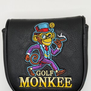 May include: A black leather-like golf putter headcover featuring an embroidered graphic of a monkey wearing a top hat, monocle, and suit, holding a cigar. The text GOLF MONKEE is embroidered in yellow below the monkey graphic on the front.