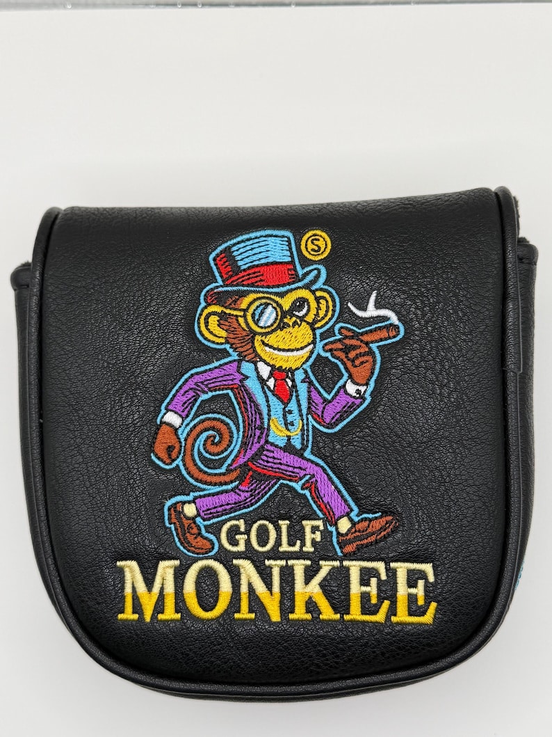 May include: A black leather-like golf putter headcover featuring an embroidered graphic of a monkey wearing a top hat, monocle, and suit, holding a cigar. The text GOLF MONKEE is embroidered in yellow below the monkey graphic on the front.