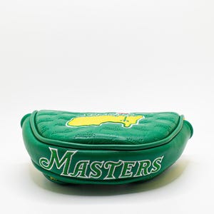 May include: A green golf putter head cover with the word "MASTERS" in white script. The cover features a yellow and green logo and a quilted pattern. The cover is made of a leather-like material.