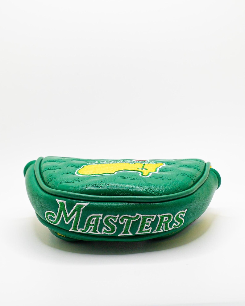 May include: A green golf putter head cover with the word "MASTERS" in white script. The cover features a yellow and green logo and a quilted pattern. The cover is made of a leather-like material.