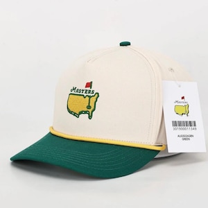 Può includere: Cappellino da baseball color crema e verde con il logo Masters ricamato sul davanti. Il cappellino ha una visiera verde e un dettaglio in corda gialla. Un'etichetta con il logo e le informazioni sul prodotto è attaccata.