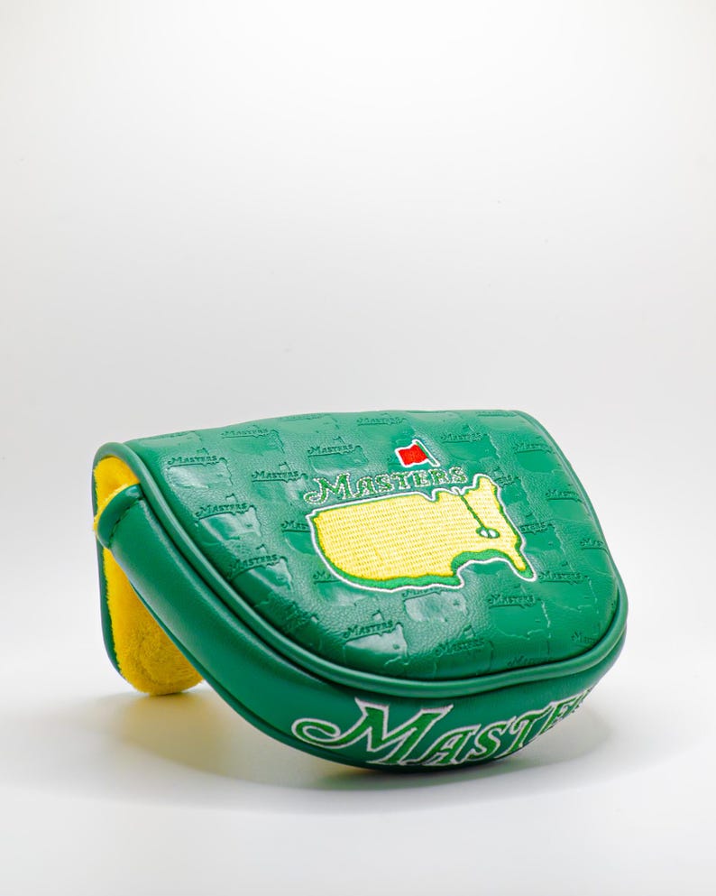 May include: Green golf club head cover with the word "Masters" in white script. The cover features a map of the United States with a red flag and the word "Masters" in green. The interior is yellow.