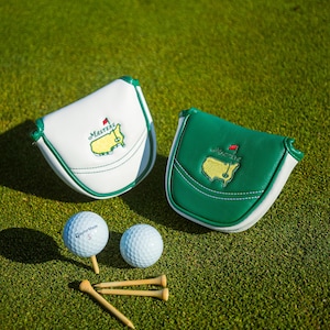 Puede incluir: Dos fundas para putter de golf, una blanca y otra verde, con el logotipo y el texto de Masters. Dos pelotas de golf y tees de madera también son visibles en un campo de golf verde.