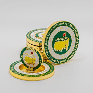 May include: Gold-colored golf ball markers with green and white accents. The markers feature the Masters logo, a map of Augusta, and the text "Masters" and "Augusta". The markers are stacked and arranged on a white surface.