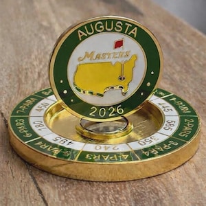Peut inclure: Un marqueur de balle de golf doré avec le texte "AUGUSTA MASTERS 2026" et une carte des États-Unis. Le marqueur est posé sur une base dorée avec des informations sur la distance et le par.