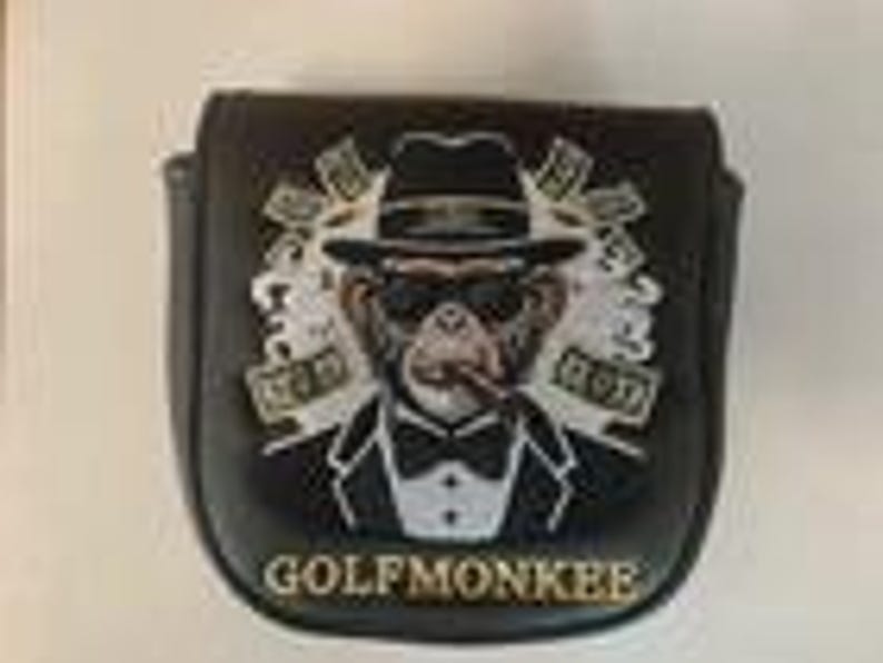 May include: A black golf putter headcover featuring an embroidered graphic of a monkey wearing a tuxedo, fedora, and sunglasses while smoking a cigar. The design includes white smoke accents and the text GOLFMONKEE printed in gold at the bottom.
