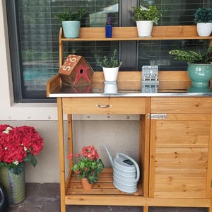May include: A wooden potting bench with a shelf, drawer, and cabinet. It holds potted plants, a watering can, and a birdhouse. The bench is made of light-colored wood and has a metal countertop.