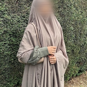 Può includere: Un'abaya color tortora caratterizzata da una silhouette ampia e fluida con copricapo abbinato. L'indumento presenta maniche lunghe con polsini arricciati verde oliva e una borsa a tracolla color tortora coordinata portata sulla spalla.