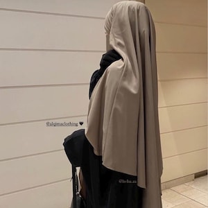 Könnte beinhalten: Eine Person trägt ein schwarzes Kleid und einen beigefarbenen Hijab. Der Hijab bedeckt Kopf und Schultern. Eine schwarze Handtasche wird in der linken Hand gehalten. Das Bild enthält den Text "@alqimaclothing" und "@hvba_ax".