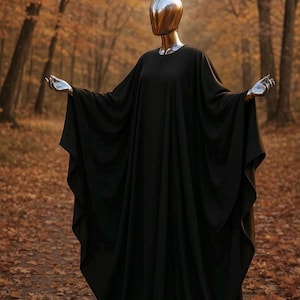 May include: A full-length black robe with wide, flowing sleeves worn by a mannequin with a smooth, reflective silver head and matching silver hands. The garment features a simple round neckline and drapes loosely to the ground in soft folds.