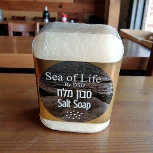 May include: A rectangular bar of salt soap, wrapped in clear plastic. The label features the text "Sea of Life" above Hebrew text and the words "Salt Soap" on a brown and white background. The soap sits on a wooden table.