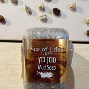 May include: A bar of Sea of Life Mud Soap in clear wrapping with a gold label. The label includes the brand name, the words "Mud Soap", and text in Hebrew. Coffee beans are scattered in the background.